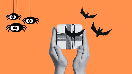 Hands holding a spooky Halloween gift box wrapped with string and decorated with bats and hanging spiders on an orange background. Perfect spooky basket gift idea for Halloween.